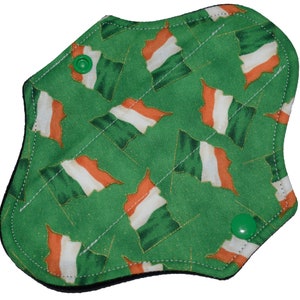 Puede incluir: Compresa menstrual de tela reutilizable con fondo verde y un estampado de banderas irlandesas en naranja, blanco y verde. La compresa tiene forma curva y se asegura con dos broches verdes.