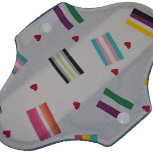 Może przedstawiać: Wielorazowy materiałowy podkład menstruacyjny z białym tłem. Podkład ma wzór kolorowych pasków i małych czerwonych serc. Widoczne są dwa białe zatrzaski. Krawędzie są jasnoniebieskie.