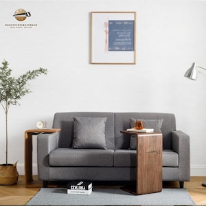 Pode incluir: Um sofá de tecido cinza com duas almofadas e uma mesa auxiliar de madeira com um design curvo. A mesa está ao lado do sofá e tem um livro e um copo em cima. O sofá está em frente a uma parede branca com uma imagem emoldurada pendurada nela.