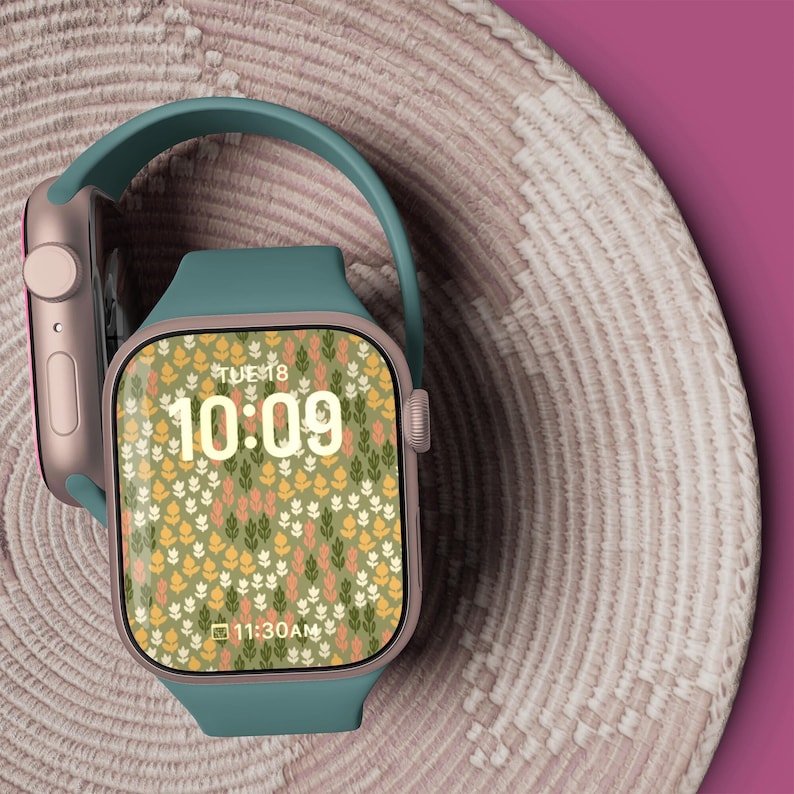 May include: A teal smartwatch with a rose gold case and a floral patterned watch face. The watch face displays the time as 10:09 and the date as TUE 18. The watch is on a woven surface.