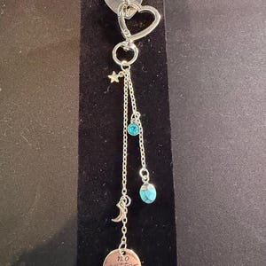 May include: Silver keychain with a heart-shaped clasp and a dangling chain. The chain features a star, a blue gem, a crescent moon, and a round charm with the words "no matter where". A small heart-shaped charm is at the top.