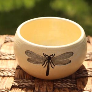 May include: A round, cream-colored ceramic bowl with a dragonfly design. The dragonfly is depicted in black and is centered on the bowl's exterior. The interior of the bowl is a light yellow color. The bowl is sitting on a woven surface.