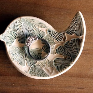 May include: A ceramic ring dish in the shape of a bird, with a silver ring resting inside. The dish is white with a green leaf pattern. The ring dish is on a wooden surface.