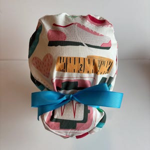 May include: A white scrub cap with a blue bow. The cap features a pattern of pink running shoes, measuring tapes, and hearts. The design also includes a pink and black EKG-like graphic.