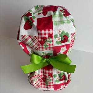 May include: A scrub cap with a patchwork design featuring red strawberries, white flowers, and green leaves on a white background. The cap has a green bow at the base. The fabric has a mix of red and white gingham and solid red squares.