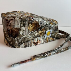 May include: Camouflage print scrub cap with a smiley face patch. The cap features a realistic pattern of trees, leaves, and deer. It has long ties for securing the cap. The cap is made of a soft, durable fabric.