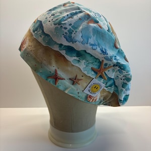 May include: A scrub cap featuring a beach-themed print with blue ocean waves, tan sand, and orange starfish. The fabric has a white tag with a yellow smiley face and the text Jolly Molly's Scrubs. The cap has a gathered design and pleated top.