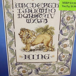 May include: Cross stitch pattern featuring a lion with a crown, surrounded by leafy vines and the word "KING". The alphabet is displayed above the lion in a decorative font.