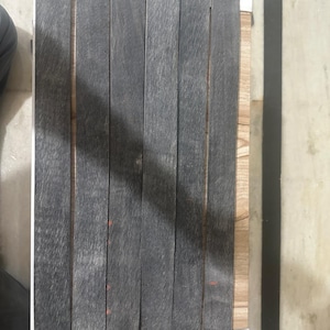 Puede incluir: Un conjunto de seis tablones de madera grises con la parte superior redondeada, dispuestos verticalmente. Los tablones están sujetos por un marco de madera con un borde blanco. La veta de la madera es visible.