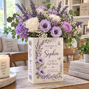May include: A rectangular white ceramic vase shaped like a book, featuring purple floral illustrations, the text "Chapter 70", "Sophia", "A Life in Full Bloom", and "Celebrating love, joy, and beautiful memories", filled with white roses and purple lavender.