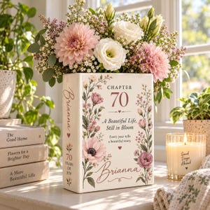 May include: A white ceramic book-shaped vase featuring pink floral illustrations and the text Chapter 70, A Beautiful Life, Still in Bloom, Every year tells a beautiful story, and the name Brianna, filled with a bouquet of pink and white flowers.