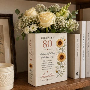 May include: A rectangular ceramic vase shaped like a book, featuring the text "CHAPTER 80", "A beautiful life, still blooming", and the name "Amelia". It is decorated with sunflower illustrations and filled with a bouquet of white roses and greenery.
