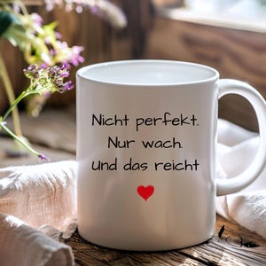Könnte beinhalten: Weiße Keramiktasse mit schwarzem Text, der "Nicht perfekt. Nur wach. Und das reicht" lautet, und einem kleinen roten Herzen.