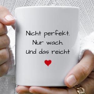 Könnte beinhalten: Weiße Keramiktasse mit schwarzem Text, der "Nicht perfekt. Nur wach. Und das reicht" lautet, und einem kleinen roten Herzen.