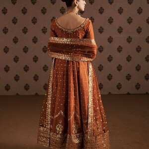 May include: A rust-colored formal gown with gold embellishments. The long-sleeved dress features intricate gold embroidery along the neckline, sleeves, and hem. The gown is worn with a matching shawl and gold jewelry.