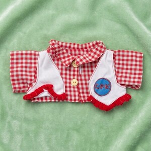 Puede incluir: Un conjunto para muñeca que incluye una camisa de cuadros rojos y blancos y un chaleco blanco. El chaleco tiene ribetes rojos y un parche azul con las letras "CPK". La camisa tiene dos botones de madera.