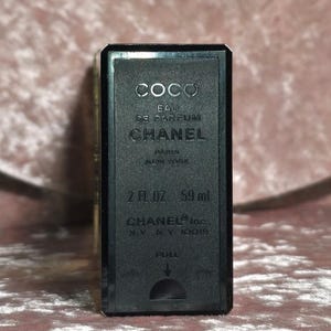 May include: A black rectangular perfume bottle with the text "COCO EAU DE PARFUM CHANEL PARIS NEW YORK" printed in white. The bottle contains 2 fl oz, which is equivalent to 59 ml. The bottom of the bottle reads "CHANEL Inc. N.Y., N.Y. 10019".
