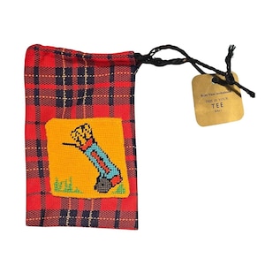 May include: A red and navy blue plaid drawstring pouch featuring a yellow square patch with a cross-stitched golf bag design. A tan rectangular tag is attached to the black drawstring, printed with the text: Ruby View Invitational THIS IS YOUR TEE BAG.