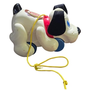 Può includere: Un giocattolo vintage a forma di cane bianco con orecchie e macchie nere. Il cane ha un collare rosso e ruote blu. Una corda gialla è attaccata per tirare. Il giocattolo ha la scritta "PLAYSKOOL".