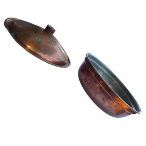 May include: A round copper pot with a matching lid. The exterior of the pot and lid features a warm, metallic copper finish with visible signs of oxidation, including small green spots on the lid. The interior of the pot has a silver-toned lining.