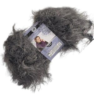 May include: A ball of grey, fluffy yarn with a label featuring a woman wearing a purple top. The yarn appears soft and textured, suitable for knitting or crocheting projects. The label includes the text "FREE GRATIS GRATUIT".
