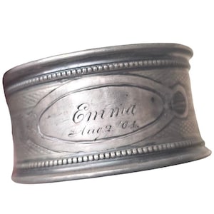 May include: A silver metal napkin ring, engraved with the name "Emma" and the date "Aug 2 '64". The ring features a beaded border and an oval design. The ring is likely made of pewter.