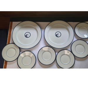 May include: A collection of white ceramic plates and saucers. The plates have a navy blue trim and a navy blue emblem in the center. The saucers also have a navy blue trim. The plates and saucers are arranged on a white surface.