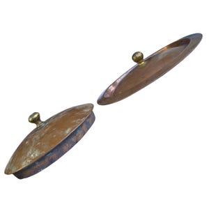 May include: Two round copper lids with a weathered, metallic finish and a polished brass knob handle centered on each. The lids feature a slightly domed shape with a raised rim, showing signs of oxidation and natural patina on the copper surface.