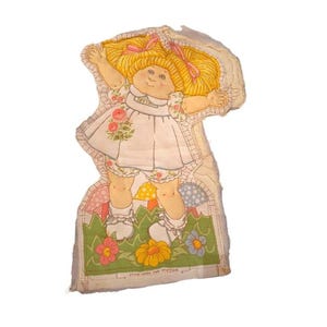 Pode incluir: Painel de boneca de tecido vintage com uma menina sorridente com cabelo loiro e laços rosa. Ela usa um vestido branco com detalhes florais e bloomers. O painel inclui uma área gramada verde com flores e cogumelos. O texto na parte inferior diz "LEAVE OPEN FOR STUFFING."