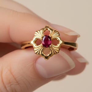 May include: A gold ring featuring a floral design with a central oval ruby. The ring's band is slender, and the setting has an openwork pattern. The ruby is a deep red colour, and the ring has a polished finish.