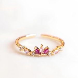 Puede incluir: Un anillo dorado con un grupo de piedras preciosas rosas y transparentes. El anillo presenta un diseño delicado con una fila de pequeñas piedras brillantes. Las piedras están dispuestas en un patrón floral o de corona.