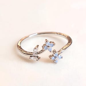 May include: A silver ring with a floral design. The ring features small, light blue flower-shaped stones and clear accents. The ring is set against a soft, off-white background, highlighting its delicate details.