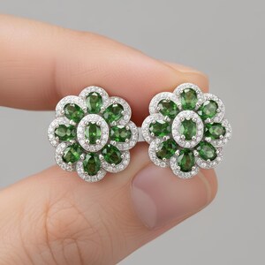 May include: A pair of flower-shaped stud earrings featuring a central oval-cut green gemstone surrounded by a halo of smaller oval-cut green gemstones and a border of tiny clear white stones set in a bright silver-toned metal frame.