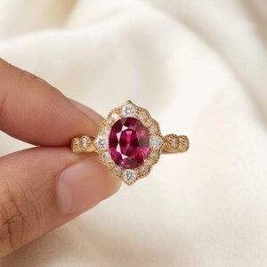 Può includere: Un anello in oro con una gemma centrale di forma ovale color rosa intenso, circondata da un'aureola smerlata di piccole pietre bianche. La fascia è realizzata in oro giallo e presenta un design in metallo testurizzato e decorativo.