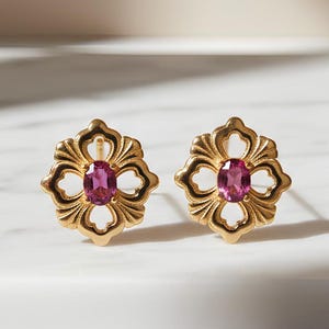 May include: Two gold-coloured stud earrings, each with a floral design and an oval, pink gemstone centre. The earrings are presented on a white surface, with soft lighting accentuating their intricate details and elegant style.