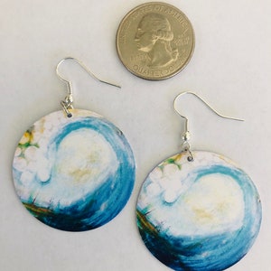 May include: A pair of round earrings with a blue and white watercolor painting of a wave. The earrings are silver and have a hook closure.