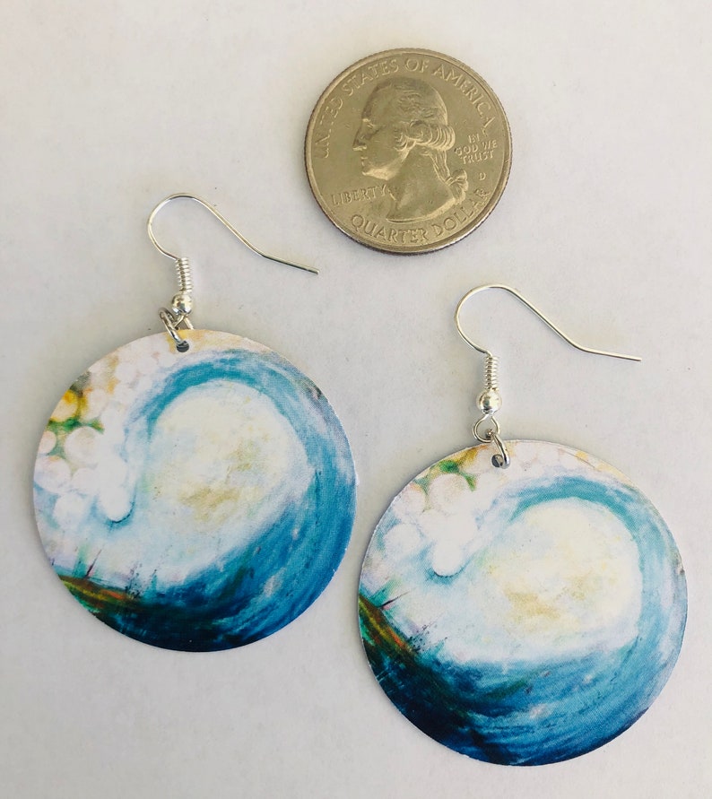 May include: A pair of round earrings with a blue and white watercolor painting of a wave. The earrings are silver and have a hook closure.