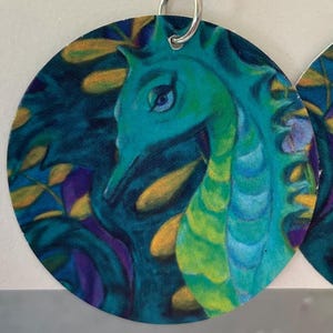 May include: Round earrings featuring a teal seahorse design. The seahorse is painted with shades of teal, green, and yellow, set against a dark blue background with yellow and purple leaf accents. The earrings have a silver hoop.