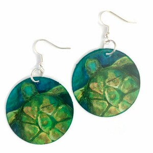 May include: A pair of round earrings featuring a painted sea turtle design. The earrings have a silver hook and are primarily green and blue, with a detailed turtle illustration. The earrings are set against a white background.