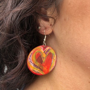 May include: A pair of dangle earrings with a round, red, yellow, green, and purple heart design. The heart has a white outline and the words "Just Love" are written on the side of the heart.