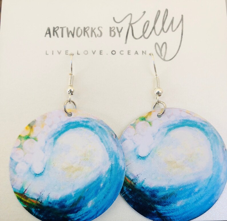 May include: A pair of blue and white wave earrings with silver hooks. The earrings are round and feature a painted design of a wave crashing. The text "ARTWORKS BY Kelly LIVE.LOVE.OCEAN." is visible in the background.