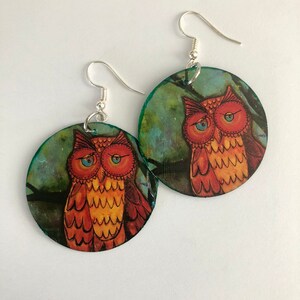 May include: A pair of round earrings featuring an owl design. The owls are depicted with orange and yellow feathers, set against a teal and green background. The earrings have silver-colored hooks.