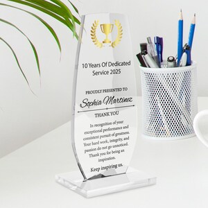 May include: A clear acrylic award with gold text and a gold trophy graphic. The text reads "10 Years Of Dedicated Service 2025" and "Sophia Martinez". A white pen holder with pens and pencils is to the right.