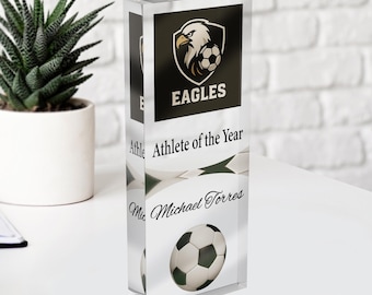 Può includere: Un premio in acrilico trasparente a tema calcio. Il premio presenta il testo "EAGLES" e "Athlete of the Year", insieme a una grafica di pallone da calcio e al nome "Michael Torres". Il premio è rettangolare e si erge in posizione verticale.