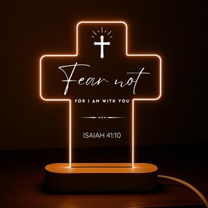 Könnte beinhalten: Ein leuchtendes, kreuzförmiges LED-Nachtlicht. Das Licht leuchtet orange und zeigt die Worte "Fear not" und "For I am with you" sowie die Bibelstelle Jesaja 41,10. Es steht auf einem Holzsockel.