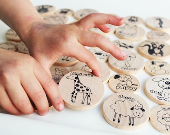 May include: Wooden animal matching game with black illustrations and text. The game features a giraffe, puppy, sheep, and other animals. A child's hands are interacting with the game pieces.