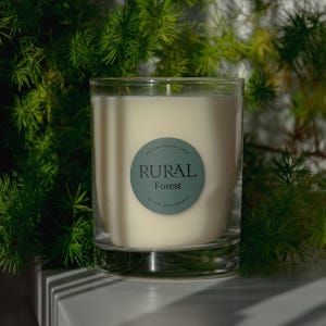 May include: A clear glass candle jar with a white candle inside. The jar has a green label with the text "ESTABLISHED 2009 RURAL Forest IN THE FOOTSTEPS".