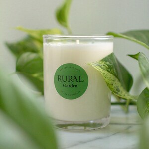 May include: A white candle in a clear glass jar with a green label that reads "ESTABLISHED 2019 RURAL Garden IN THE KOOTENAYS".
