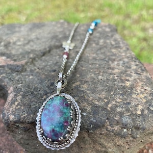 May include: A silver chain necklace with a green and red oval gemstone pendant. The pendant has a silver filigree design around the stone.