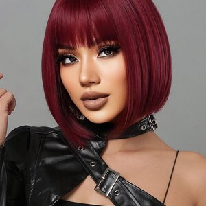A woman with vibrant red hair styled in a short bob, wearing a black leather jacket and posing against a gray background.